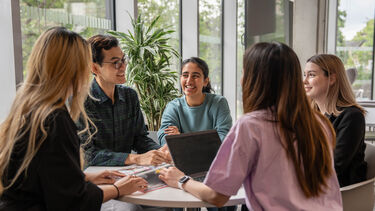 A group of postgraduate students sat around a table smiling and chatting