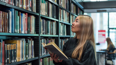 Student holding a book stood in front of bookshelf looking up at the books