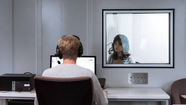one student in the sound booth speaking into a microphone as another student analyses the soundwaves outside of the booth