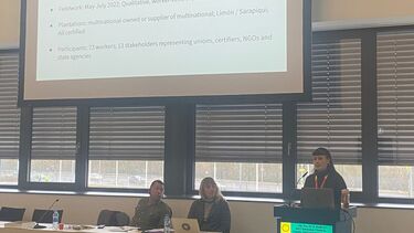 REmi stands at a lecturn in front of a huge projector screen that displays information about her methodology