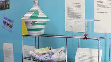 A museum display featuring items accompanied by text descriptions. There is a white and green woven basket in the shape of an african style house, and a pencil case which has images depicting Bermuda. 