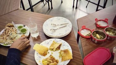 Alt text: People sharing food with plates of stuffed savoury crepes or dosas, parathas or flatbreads, and containers of curry in a room with tables and chairs.