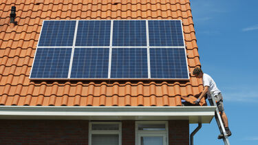 Am image of a man up a ladder installing some solar panels on the roof of a typical semi-detached house.