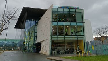 A view of MIMA, photographed by Grace. A glass and brickwork building stands against a grey sky. On the left side of the building, yellow lettering spells out MIMA; on the right, two people in yellow high-vis walk along the middle of three floors. 