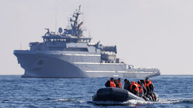 A small boat crossing the sea, in the background a much larger military or security type vessel can be seen.