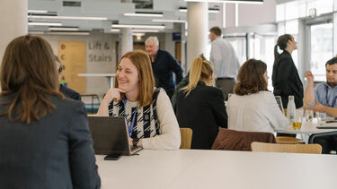 People sat at tables in a lobby, with a woman smiling in front of a laptop in the foreground