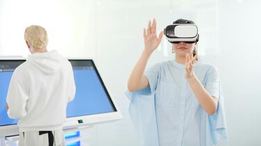 Woman in hospital gown uses a VR headset, while a person uses a screen in the background to control the headset.