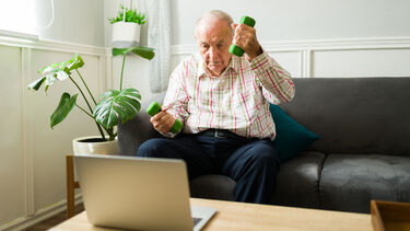 An elderly man using weights whilst watching something on a laptop in his living room.