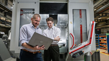 Two men stand in front of a manufacturing machining centre looking into a laptop.