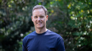 Dan Walker smiling against a backdrop of trees in Sheffield Botanical Gardens, head and shoulders shot with mid-length blond hair and navy blue t-shirt