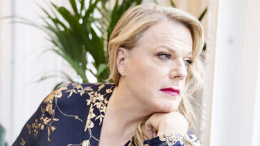 Suzy Eddie Izzard facing sideways to look out of a window, resting her chin on her hand, wearing long blone hair and a blue and gold dress
