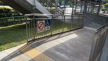 A wide ramp with tactile floor markers at an accessible overhead bridge along the Ayer Rajah Expressway in Singapore. The ramp leads to a lift that goes up and down the overhead bridge. Attached to the ramp's railing are a sign prohibiting riding, and another sign showing a wheelchair-user with an arrow pointing down the ramp. In the image background are the staircase of the overhead bridge and a bus stop.  