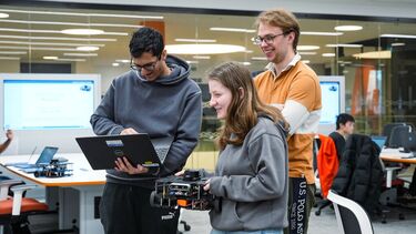 Three students are standing together in computer room, one student is holding a laptop and another one is holding a robot, the third one is standing and smiling.