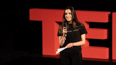 Maria stood in front of a backdrop that holds the bold, red, TEDx logo at the event she arranged at the University of Sheffield. She wears a black tshirt, has long brown hair and holds a mic whilst smiling and speaking to the crowd.
