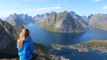 A female sits on a rock with her back to the camera looking out at a beautiful vista of mountains and water.