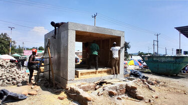 Two builders standing inside a half-finished sustainable cold room with one builder working on the roof. 