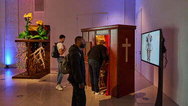  Three people interact with two distinct art installations in a gallery setting. One person is peering into a wooden booth with red curtains, while two others stand nearby. To the left, there's a sculpture of a yellow flower on a wooden base, and on the right, a large screen displays an image of an elephant. The room is lit with soft blue and pink light.