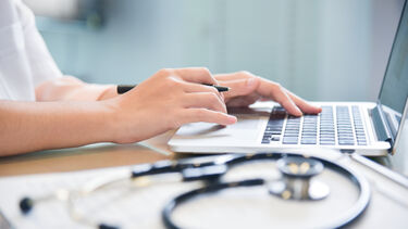 A person works at a laptop with a stethoscope visible in the foreground.