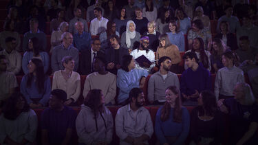 Image shows staff and students gathered in a lecture theatre with two of them picked out by a spotlight looking at a book. The book represents the University of Sheffield's shared ambition.