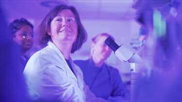 Heather is in a lab coat looking up from a microscope, there is a purple colour filter on the image