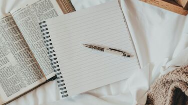 A bed with the covers pulled back and a notepad open at a page to write. It is next to an open book.  There is a cup of coffee on a tray. 