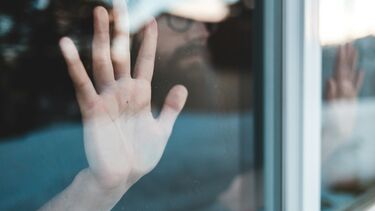 A man wearing glasses looks out of a window with his hand pressed up against the glass. 