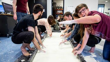 General Engineering students are doing a Newtonian fluid experiment in the lab. They are squatting beside the container of plaster. A male student is smiling, showing his hand with plaster liquid on it.