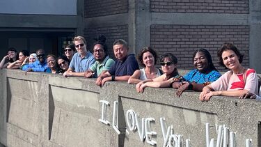 A group of people stand in a line on a bridge and smile towards the camera. Below them is a sign on the bridge that reads "I love you"