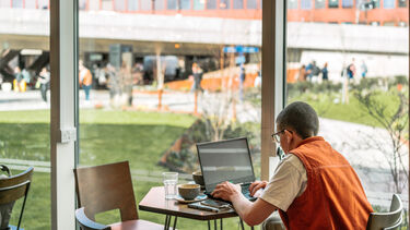 A person is seated at a table in front of a full-length window, using a laptop