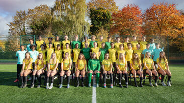 A student sports team is arranged in three rows in an outdoor setting, looking at the camera and smiling. Behind them is a row of trees.