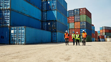 Mid distance view of four people wearing reflective vests and hardhats amidst stacks of cargo containers.