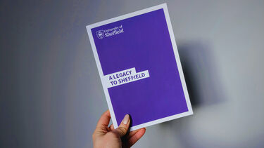 Image of a hand holding up a purple and white Sheffield-branded brochure titled "A legacy to Sheffield" against a plain grey background