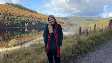 Sophie wears a green jumper and red scarf, and stands in a Peak District landscape