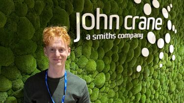 A young man stands in front of a green wall with the text 'John Crane'