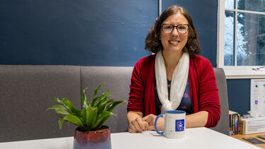 Katherine has a short curly bob and wears a red cardigan. She sits on a grey chair in front of a blue wall.