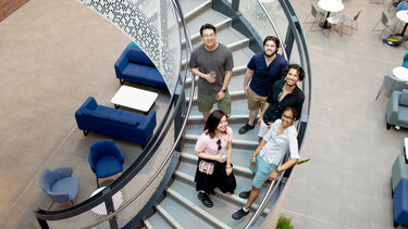 A group of five students stand on an internal winding staircase and look up at the camera, smiling.