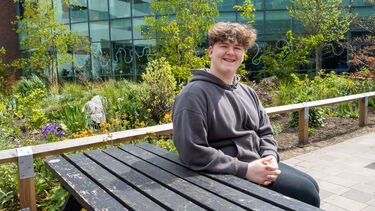 Alfie wears a grey hoodie and sits at a picnic bench. The SU and lots of plants are behind him.