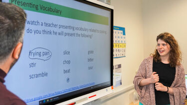 A female student stands in the background of the image and looks to her left where there is a screen displaying a selection of English words. She holds a pen in her hand and is mid-speech, presenting.