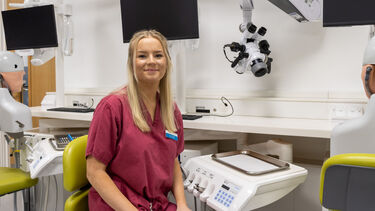Lucy has blonde hair and wears red scrubs. She sits in a chair with dental phantom heads behind her.
