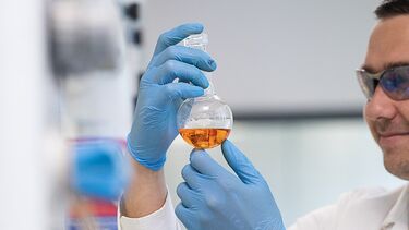 A lab worker is holding up a bottle with an orange liquid inside.