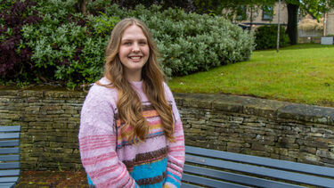 Charlotte has long blonde hair. She wears a pink stripy jumper and stands in front of some trees and bushes.