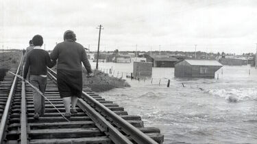 Black and white photo of two people walking on a railway line above a flooded landscape