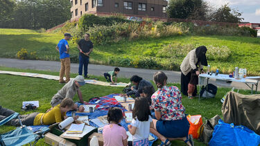 A group of people with blurred faces doing arts and crafts on the grass in front of a block of flats