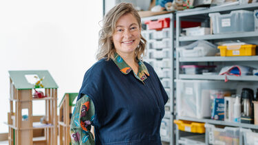 Emma Horton stood in front of shelves with lots of trays on and arts and crafts materials, there is also a wooden dolls house in background. 