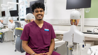 Randeep has dark curly hair, and wears burgundy dental scrubs. He sits on a chair next to a row on phantom head simulators.