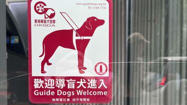  Red and white sticker from Hong Kong Guide Dogs Association on a glass door that shows a drawing of a guide dog with the caption "Guide dogs welcome". Blue and white wheelchair symbol sticker is below.