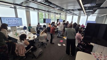 People standing and stood up in a seminar room gathered around tables, playing pictionary