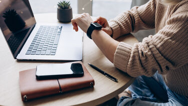 A woman sat at a desk looking at her smartwatch with a laptop and mobile phone opposite her on the table