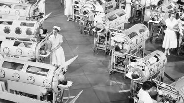 Black and white photo of hospital ward with rows of people inside iron lungs which look like large cylinders 