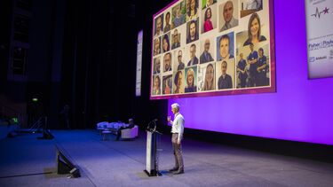 Steve Goodacre stands on a stage behind a lectern with slides of colleagues projected behind him. 
